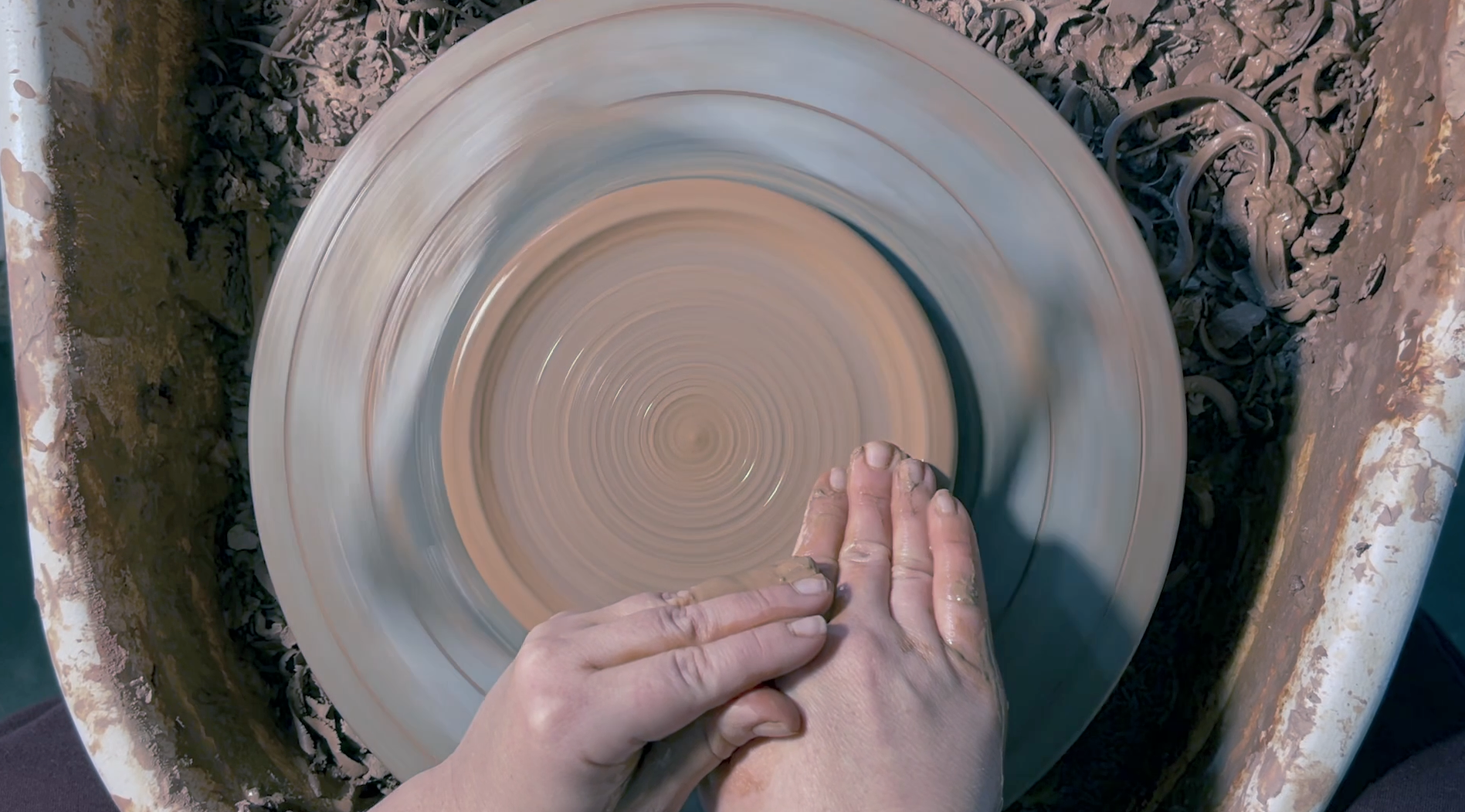 Load video: Video of a handmade ceramic vessel being made on a pottery throwing wheel. We see a lay-down angle of female hands shaping dark red, moist clay while it spins hypnotically on the wheel head making perfect spirals.